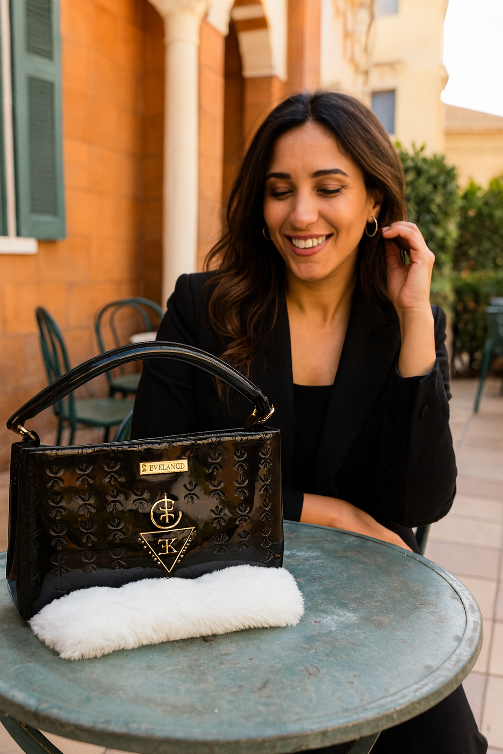 Sac TASSEDA en cuir premium noir avec logo doré 3D K.ELEGANCE posé sur une table ronde avec femme souriante en arrière-plan